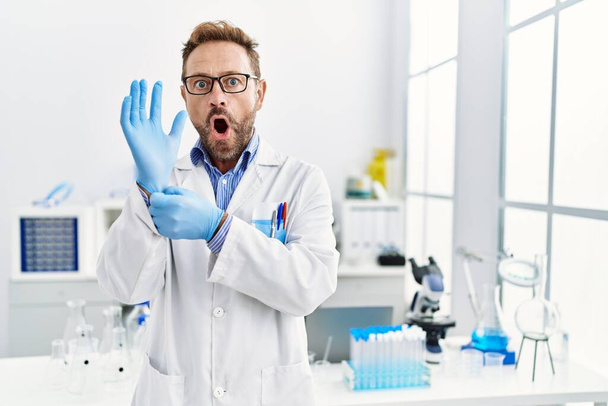 Homem de meia-idade trabalhando no laboratório de cientistas com medo e chocado com a expressão surpresa e surpreso, medo e rosto animado.  - Foto, Imagem