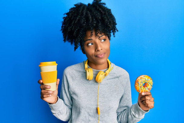 Afrikai amerikai nő afro hajjal fánkot eszik és kávét iszik mosolyogva néz oldalra, és félrenéz gondolkodás közben..  - Fotó, kép