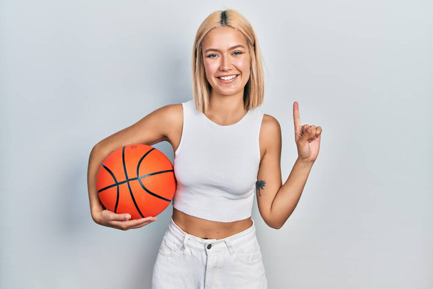 Gyönyörű szőke nő kezében kosárlabda mosolyogva egy ötlettel vagy kérdéssel ujjal mutogató boldog arc, első számú  - Fotó, kép