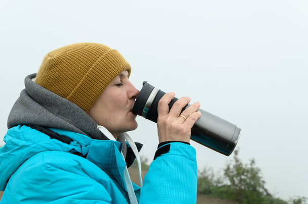 A woman in a warm jacket with a hood and a knitted hat drinks from a thermos mug. The entire background is clouded with a white milky mist. - Valokuva, kuva
