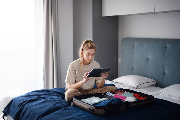 Portrait of young woman sitting on bed indoors at home, planning holiday. - Foto, afbeelding