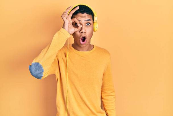 Junger afrikanisch-amerikanischer Typ, der Musik über Kopfhörer hört und dabei eine ok Geste macht, schockiert mit überraschtem Gesicht und Augen, die durch Finger schauen. Ungläubiger Ausdruck.  - Foto, Bild