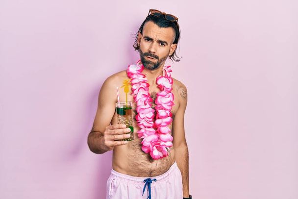 Jonge Spaanse man in badpak en Hawaïaanse lei drinkt tropische cocktail in shock gezicht, ziet er sceptisch en sarcastisch uit, verrast met open mond  - Foto, afbeelding