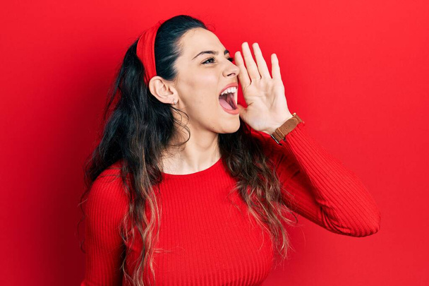 Jonge Spaanse vrouw draagt nonchalante kleren schreeuwend en schreeuwend naast elkaar met hand op mond. communicatieconcept.  - Foto, afbeelding