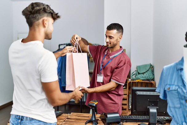 Jonge Spaanse winkelier glimlachend blij het geven van boodschappentas aan de klant in de kledingwinkel. - Foto, afbeelding