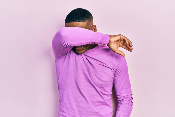 陽気で面白い腕で目を覆うカジュアルなピンクのセーターを着た若い黒人男性。視覚障害の概念.  - 写真・画像