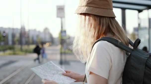 Młoda turystka z plecakiem szukająca właściwego kierunku na mapie czeka na transport publiczny na dworcu tramwajowym na zewnątrz. Podróż do Europy - Materiał filmowy, wideo