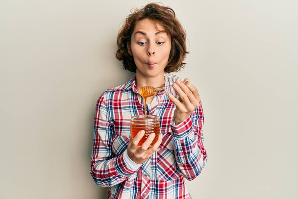 Young brunette woman holding fresh honey making fish face with mouth and squinting eyes, crazy and comical.  - Foto, imagen