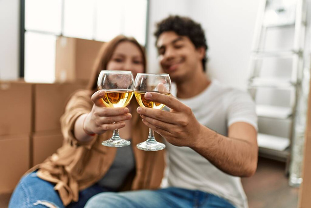 Νεαρό ζευγάρι χαμογελά ευτυχισμένη πρόποση με ένα ποτήρι κρασί στο νέο σπίτι. - Φωτογραφία, εικόνα