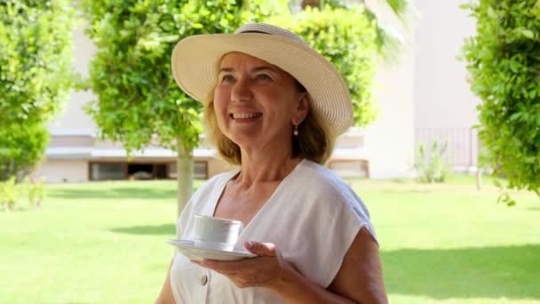 Une femme adulte souriante âgée de 50 à 55 ans dans un chapeau de paille boit une boisson chaude de café ou de thé dans une tasse blanche tôt le matin sur la terrasse de sa maison dans un jardin verdoyant par une journée ensoleillée. Senior senior - Séquence, vidéo