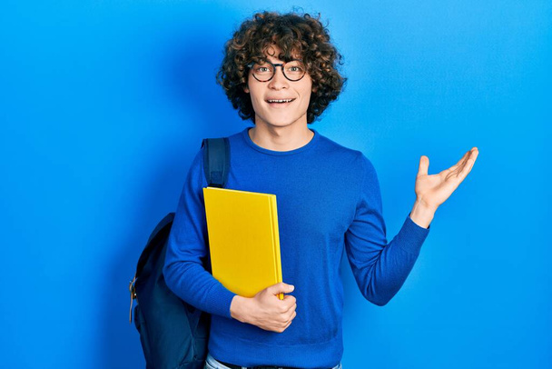 Schöner junger Mann mit Studentenrucksack und Buch in der Hand, der den Sieg mit glücklichem Lächeln und Siegerausdruck mit erhobenen Händen feiert  - Foto, Bild