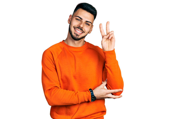 カジュアルなオレンジのセーターを着た髭を生やした若いヒスパニック系の男が勝利サインをしてカメラに顔をウィンクして微笑んだ。2番.  - 写真・画像