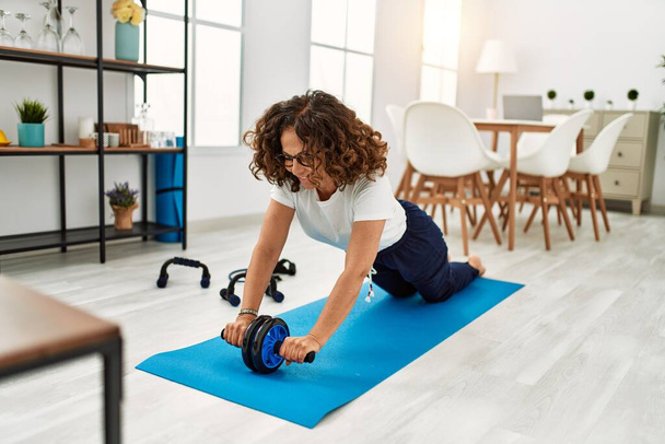 Mature hispanic woman doing exercise with abdominal wheel at the living room at home - Photo, Image