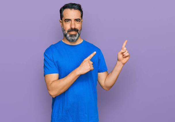 Μεσήλικας άντρας με γενειάδα φοράει μπλε μπλουζάκι χαμογελώντας και κοιτάζοντας την κάμερα να δείχνει με δύο χέρια και δάχτυλα στο πλάι.  - Φωτογραφία, εικόνα