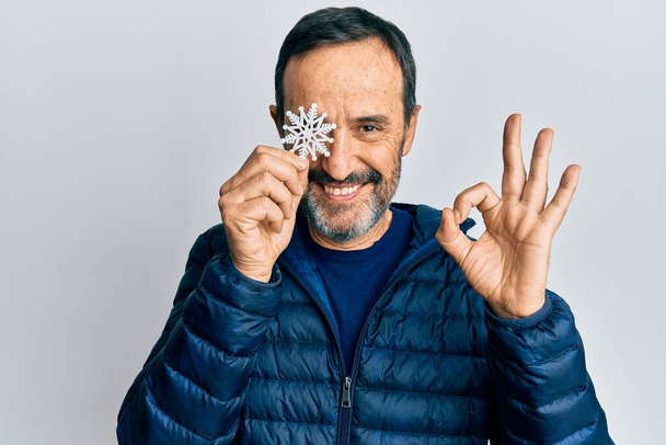 Латиноамериканец средних лет держит снежинку над глазом, делает знак "ОК" пальцами, улыбаясь дружелюбный жест отличный символ  - Фото, изображение