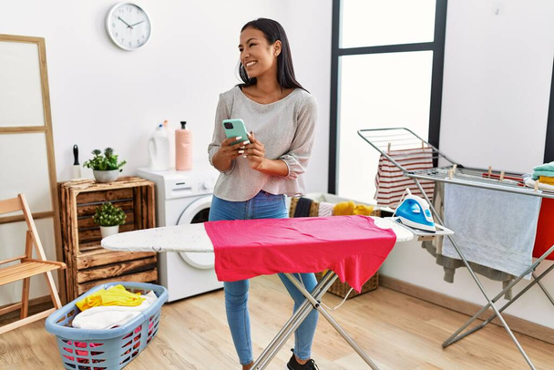 Young latin woman using smartphone ironing at laundry room - Photo, Image