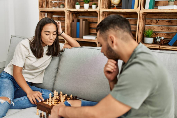 Νεαρό ζευγάρι Λατίνων χαμογελά χαρούμενος παίζοντας σκάκι στο σπίτι. - Φωτογραφία, εικόνα