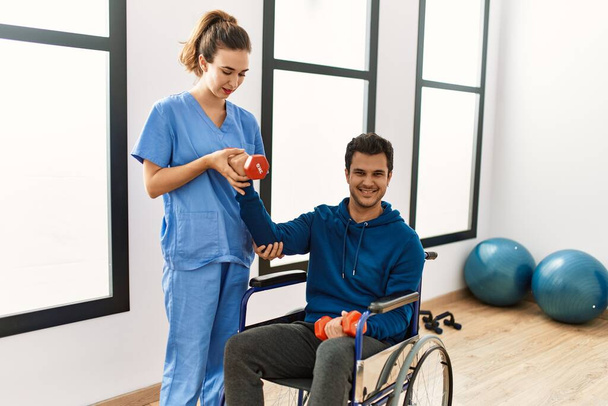 Joven discapacitado sentado en silla de ruedas haciendo ejercicio de movilidad usando pesas en la clínica. - Foto, Imagen