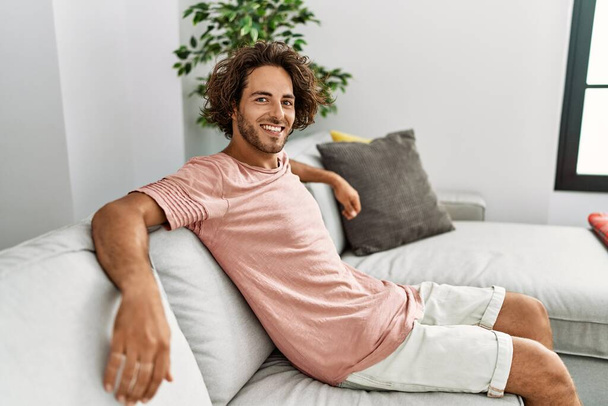 Mladý hispánec se šťastně usmívá, když sedí doma na pohovce. - Fotografie, Obrázek