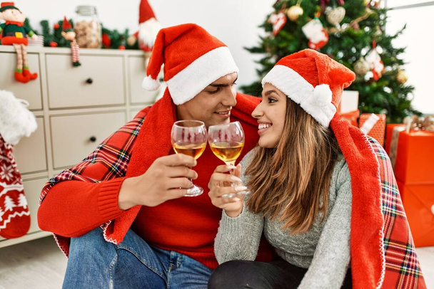 Νεαρό ζευγάρι που φοράει χριστουγεννιάτικο καπέλο καλυμμένο με κουβέρτα. Καθισμένος στο πάτωμα κάνοντας πρόποση με σαμπάνια στο σπίτι. - Φωτογραφία, εικόνα