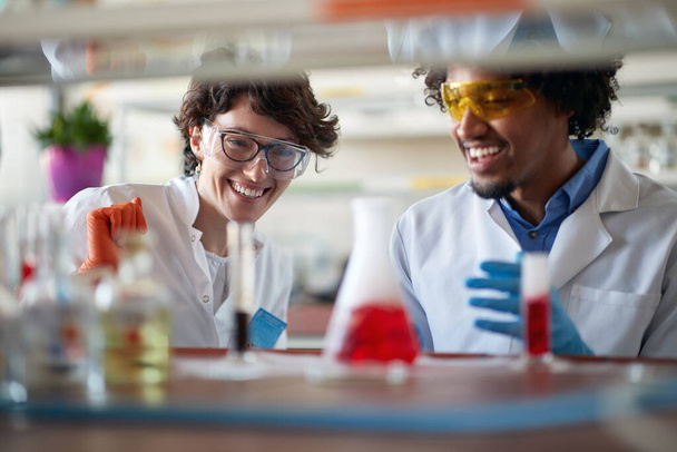 Young colleagues in a protective gear analyze colorful chemicals in a relaxed atmosphere in the university laboratory. Science, chemistry, lab, people - Photo, Image