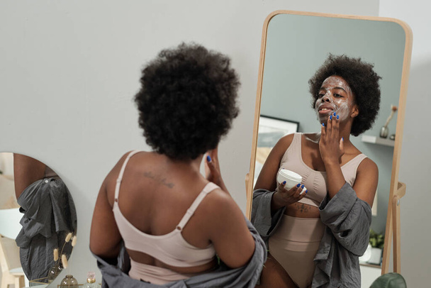 Молодая женщина наносит маску на лицо перед зеркалом - Фото, изображение