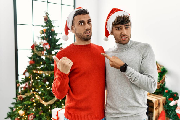 Young gay couple standing by christmas tree wearing hat pointing aside worried and nervous with forefinger, concerned and surprised expression  - Photo, Image