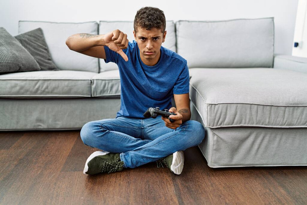 Jeune homme hispanique beau jeu vidéo assis près du canapé avec le visage en colère, signe négatif montrant détester avec les pouces vers le bas, concept de rejet  - Photo, image