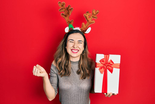 Young hispanic girl wearing deer christmas hat holding gift screaming proud, celebrating victory and success very excited with raised arm  - Foto, Imagem