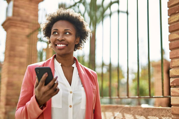 アフロの髪を持つ美しいビジネスアフリカ系アメリカ人女性は、スマートフォンを使用して街で幸せと自信を持って屋外で笑顔 - 写真・画像