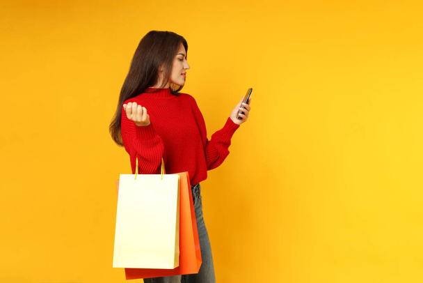 Chica atractiva con bolsas de papel y teléfono inteligente sobre fondo amarillo. - Foto, Imagen