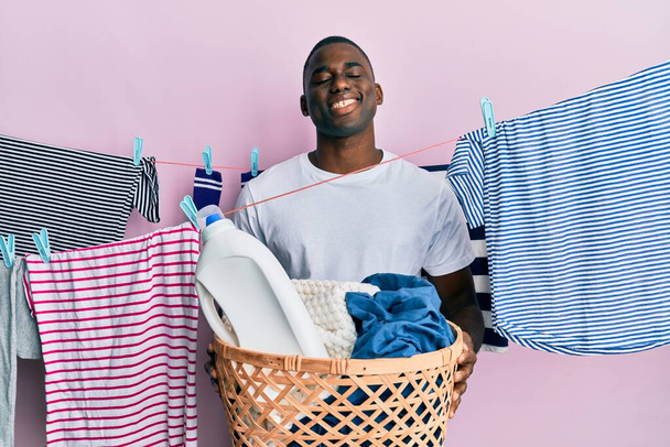 Genç Afrikalı Amerikalı adam çamaşır sepetini tutarken yüzünde mutlu ve havalı bir gülümsemeyle gülümsüyor. diş gösterme.  - Fotoğraf, Görsel