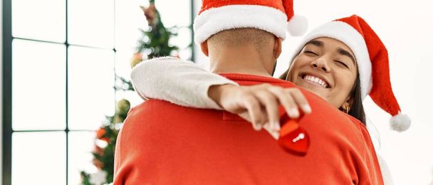 Νεαρό ζευγάρι ισπανόφωνων χαμογελά ευτυχισμένο και αγκαλιάζει κρατώντας δαχτυλίδι αρραβώνων στέκεται δίπλα στο χριστουγεννιάτικο δέντρο στο σπίτι. - Φωτογραφία, εικόνα