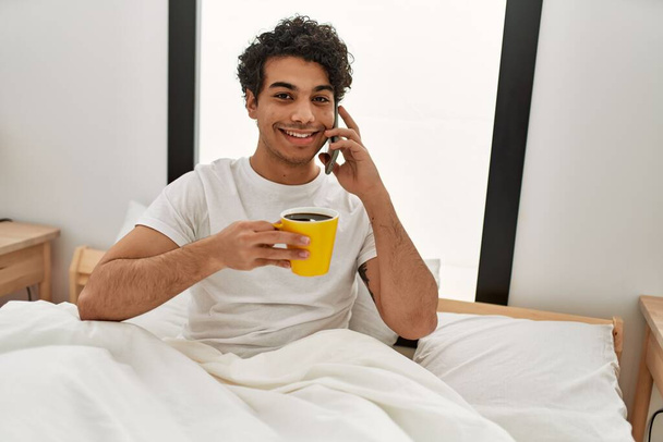 Jovem hispânico bebendo café e falando no smartphone no quarto. - Foto, Imagem
