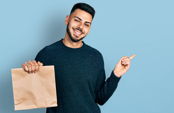 Jonge Spaanse man met baard die wegneemt papieren zak lachend vrolijk wijzend met hand en vinger naar de zijkant  - Foto, afbeelding