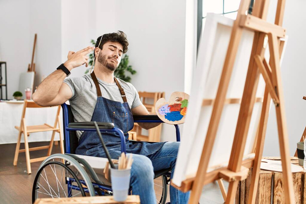 Joven hispano sentado en silla de ruedas pintando en el estudio de arte disparando y matándose apuntando de la mano y los dedos a la cabeza como arma, gesto suicida.  - Foto, Imagen