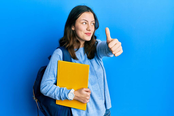 Giovane studentessa bruna che indossa uno zaino da studente facendo dei pollici verso l'alto gesto sorridente guardando di lato e fissando via pensando.  - Foto, immagini
