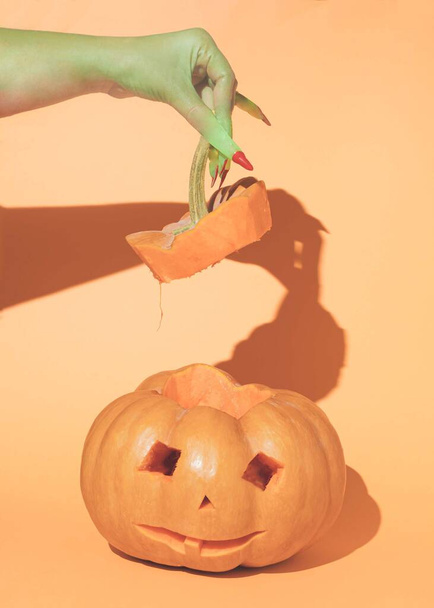 A scary witch holds a pumpkin handle on an orange background. Minimal layout - 写真・画像