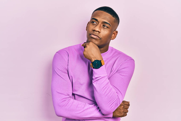 Молодий чорний чоловік в повсякденному рожевому светрі з рукою на підборідді думає про питання, стомлюючий вираз. усміхнений вдумливим обличчям. концепція сумніву
.  - Фото, зображення