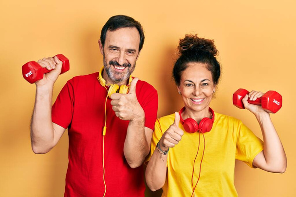 Пара латиноамериканцев среднего возраста в спортивной одежде с гантели улыбаются счастливыми и позитивными, большой палец вверх делает отлично и знак одобрения  - Фото, изображение