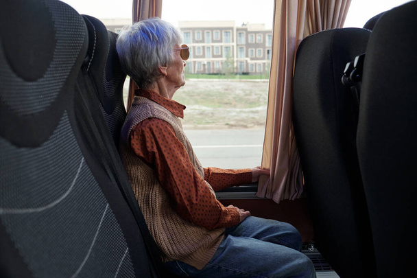 Starsza kobieta w ubraniu casualwear patrząc przez okno podczas siedzenia w autobusie - Zdjęcie, obraz