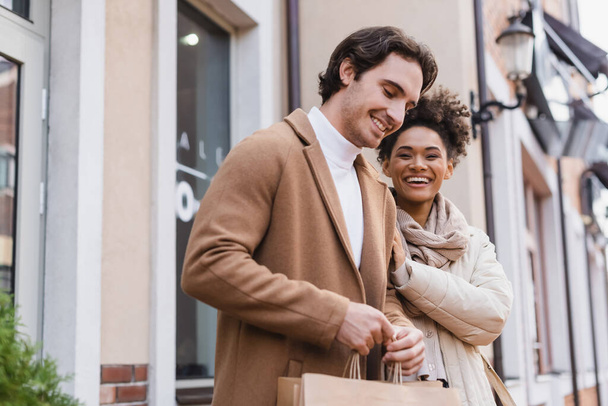 positive african american woman smiling near boyfriend holding shopping bags in mall  - Fotografie, Obrázek