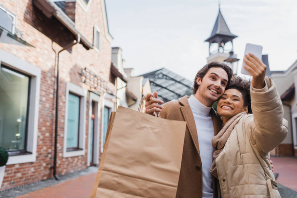 mujer afroamericana feliz tomando selfie con novio sosteniendo bolsas de compras - Foto, Imagen