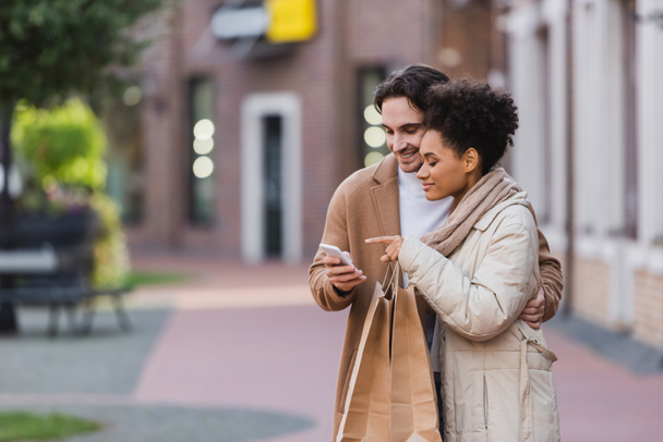 giovane donna afro-americana che punta lo smartphone vicino al fidanzato con le borse della spesa - Foto, immagini