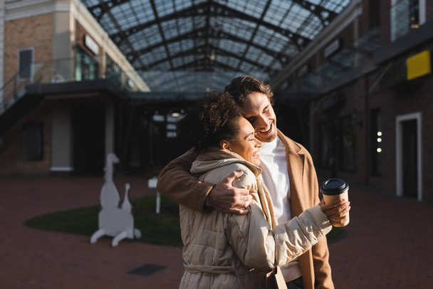 alegre hombre abrazando feliz africana americana mujer con papel taza en centro comercial  - Foto, imagen