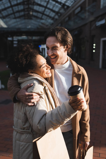 homem sorridente abraçando mulher americana africana feliz com copo de papel e saco de compras - Foto, Imagem