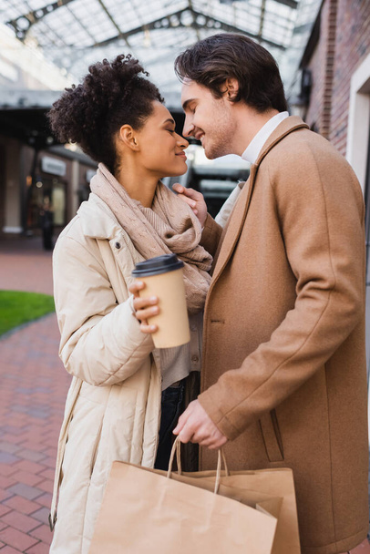 side view of happy african american woman with coffee to go near pleased boyfriend holding shopping bags - Foto, immagini