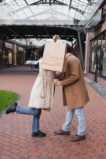 brunette african american woman holding shopping bags while covering faces of her and boyfriend in mall  - Foto, Imagem
