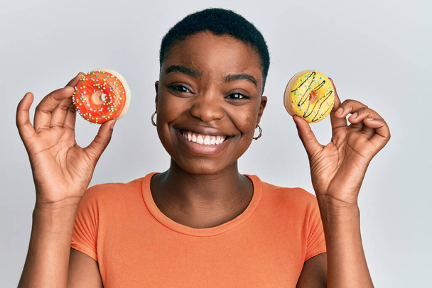 jong Afrikaans amerikaans vrouw houden smakelijke kleurrijke donuts op ogen knipperen kijken naar de camera met sexy uitdrukking, vrolijk en gelukkig gezicht.  - Foto, afbeelding