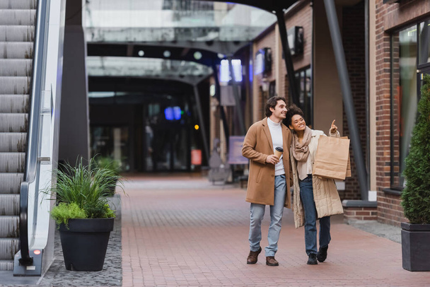 longitud completa de la mujer afroamericana sosteniendo bolsas de compras y señalando mientras camina con novio feliz en abrigo en el centro comercial  - Foto, imagen